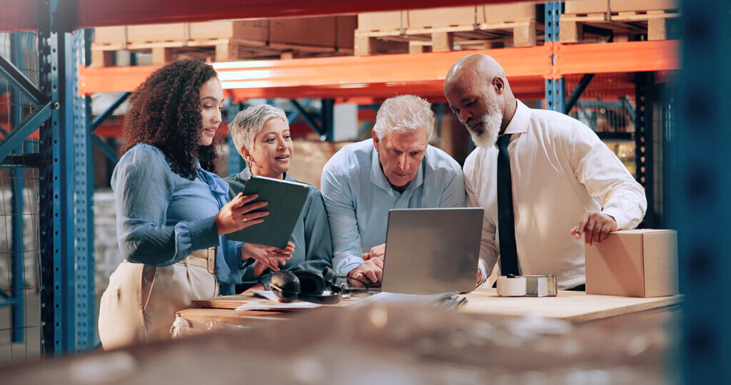 A logistics team is coordinating routed deliveries while gathered around a table in a warehouse