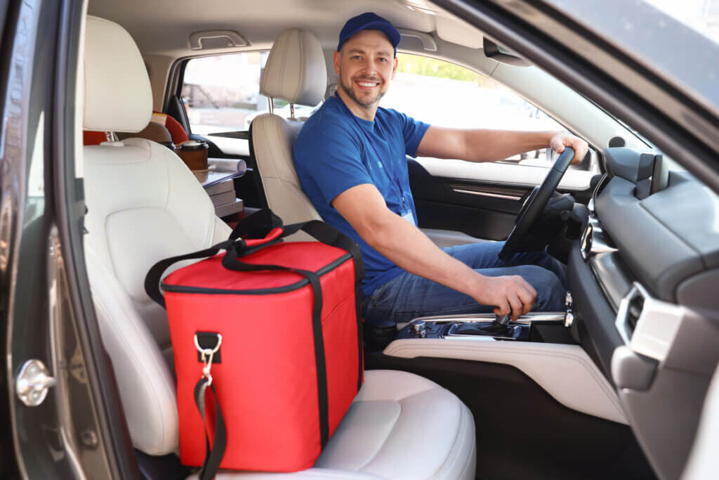 A male courier providing same day delivery services is driving with a red fabric thermo bag in the passenger seat