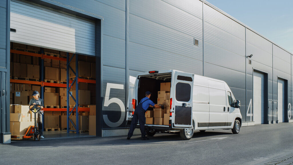 Two couriers fulfilling distribution services as they load a van with packages from a garage door at the back of a warehouse