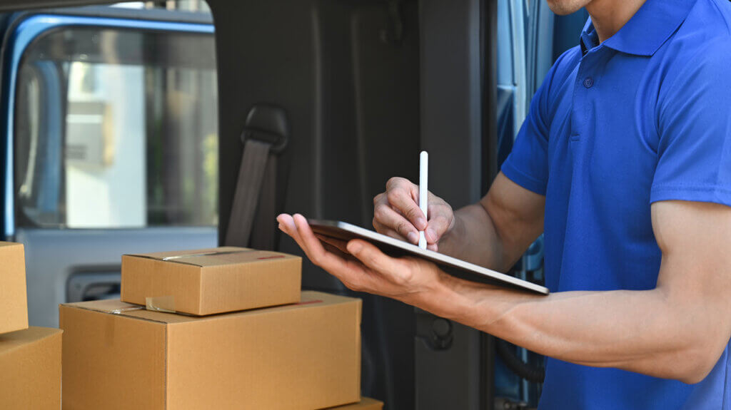 A male delivery driver is checking a list on a clipboard and reviewing stacks of packages, ensuring dependable delivery